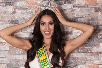 A woman smiling, adjusting a tiara on her head, wearing a sash labeled "miss brasil.