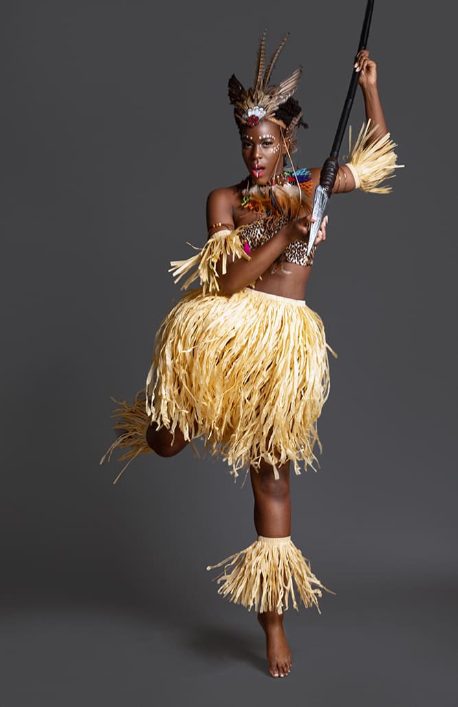 A woman in traditional african attire and headdress dances dynamically with a spear, wearing a raffia skirt.