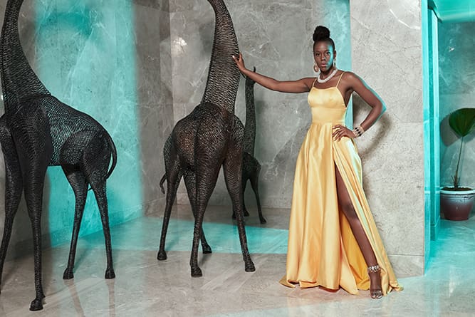 A woman in a yellow dress stands confidently next to large abstract horse sculptures in a luxurious interior.