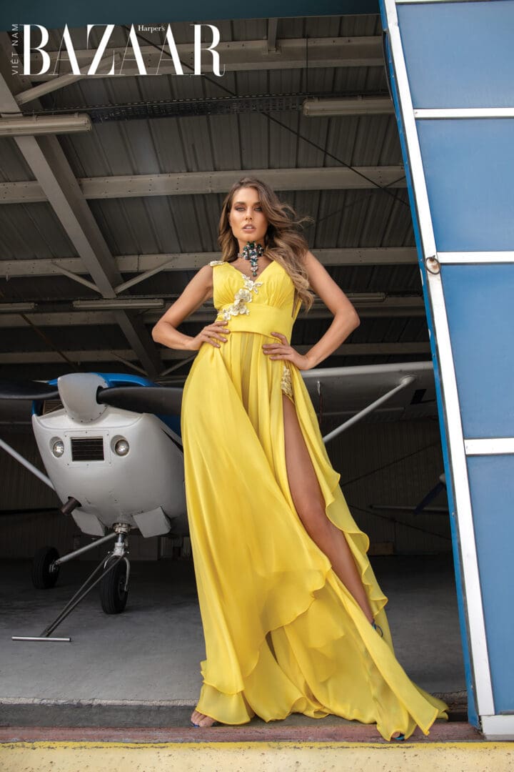 A woman in a flowing yellow dress poses by a small airplane inside a hangar, featured on the cover of harper's bazaar magazine.