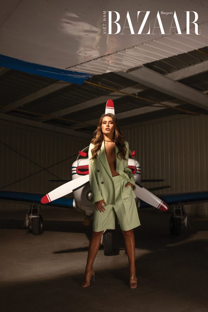 A model in a green coat poses confidently in front of a small airplane inside a hangar for a harper's bazaar photoshoot.