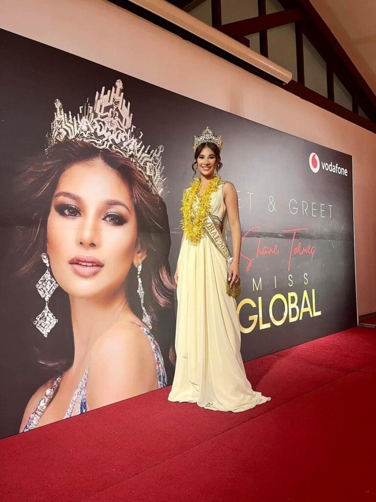 Woman in a yellow gown and crown posing in front of a promotional banner featuring her image at a meet and greet event.