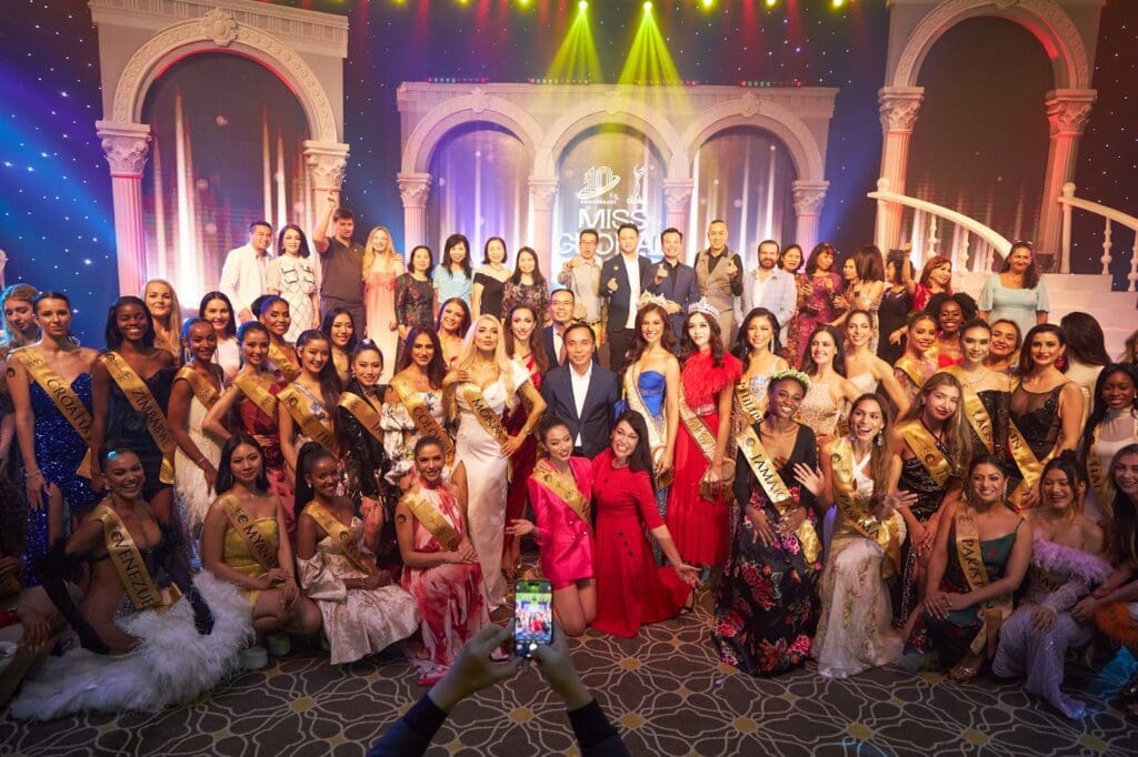 Group photo of contestants and hosts in formal wear at an international beauty pageant event with colorful stage lighting.