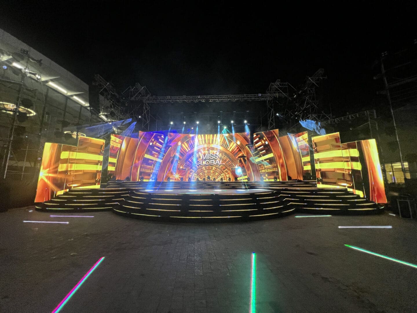 Brightly lit stage setup for miss global event at night with colorful lighting and large led screens displaying the event's title.