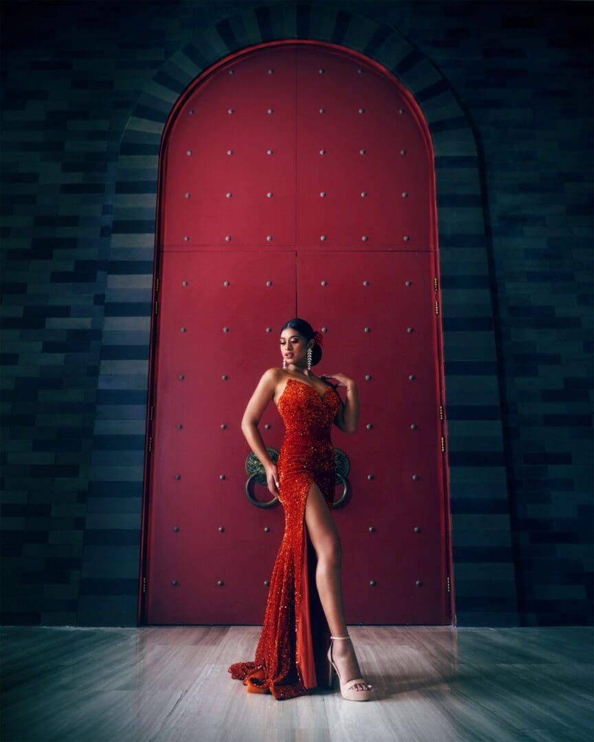 A woman in an elegant red dress with a high slit, leaning against a large red arched door, in a room with mosaic tiled walls.