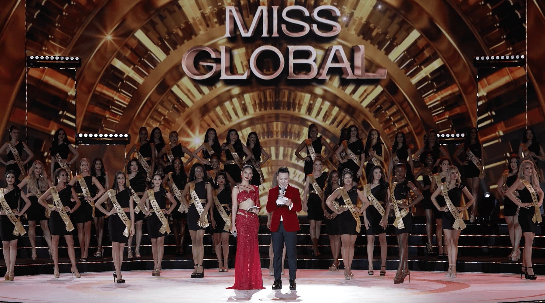 Contestants in sashes stand on stage at the miss global beauty pageant, with a host speaking into a microphone, all under a grand, illuminated archway.