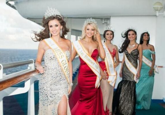 A group of women wearing sashes and crowns, representing various titles, walking in line on a cruise ship deck.